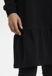 Robe noire avec ourlet à volants et manches longues. Le tissu semble doux et lisse. Une main tient la robe près du bas.