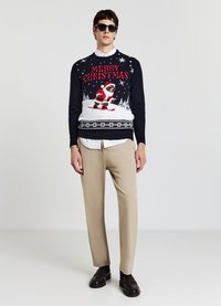 Maglione lavorato a maglia color navy con la scritta "MERRY CHRISTMAS", design di Babbo Natale che scia, fiocchi di neve bianchi e bordo con motivo. Abbinato a pantaloni beige.