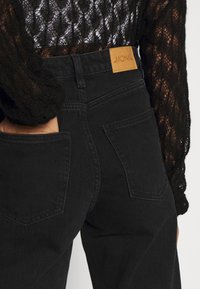 Vaqueros de mezclilla negra con una cintura alta, bolsillos traseros y una etiqueta de cuero marrón con "MONKI". La tela tiene una textura suave.