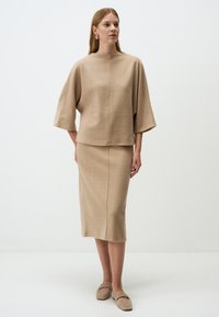 Beige gestricktes Oberteil mit weiten Ärmeln und hohem Kragen, kombiniert mit einem knielangen Rock aus demselben Material, beide mit einer glatten Textur.