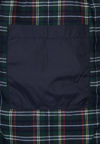 Tartan-Stoff mit einem karierten Muster in dunklem Grün, Rot, Gelb und Weiß. Verfügt über eine einfarbige schwarze rechteckige aufgesetzte Tasche in der Mitte.