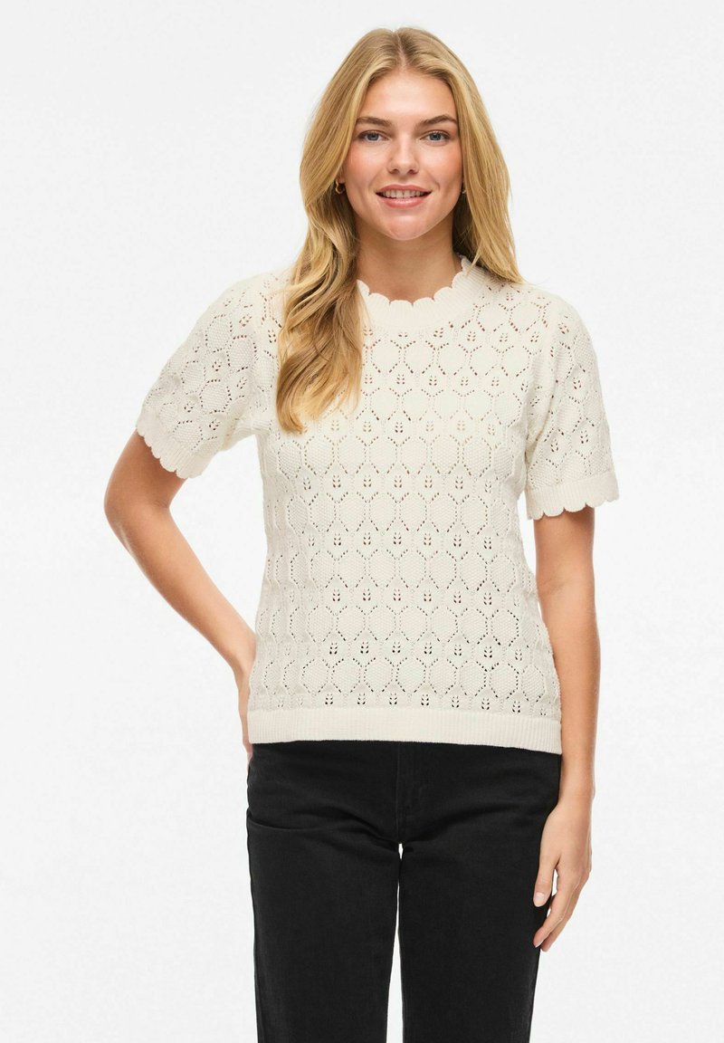 Femme blonde souriante portant un haut en maille blanc à manches courtes avec des bords festonnés et un pantalon noir, debout avec une main sur la hanche.