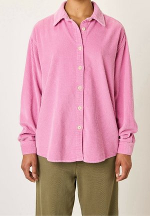 Osoba ubrana w różową sztruksową koszulę zapinaną na guziki w kolorze beżowym oraz oliwkowozielone spodnie, stojąca na jednolitym tle.