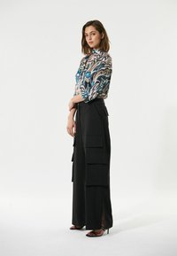 Geprinte blouse met een hoge kraag in blauw, roze en cream. Zwarte wijde broek met gelaagde zakken en zijsplitten. Zwarte sandalen.