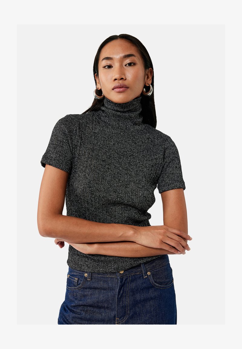 Woman with long dark hair wearing silver hoop earrings, a gray short-sleeve turtleneck sweater, and blue jeans, arms crossed at waist.