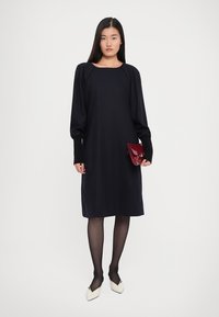 Woman wearing a black long-sleeve midi dress, sheer black tights, white pointed shoes, and holding a small red clutch purse.