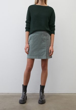 Pull en maille texturé vert foncé, jupe vert clair, bottines noires à semelles épaisses. Silhouette simple, coupe décontractée et accents minimalistes.