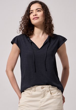 Femme aux cheveux bouclés portant un haut noir à manches courtes en V avec cordons et un pantalon beige sur fond uni.