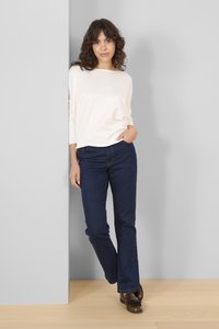 Hvid, ribbet bluse med lange ærmer i en afslappet pasform, parret med mørkeblå udringede jeans. Modellen står foran en lys grå væg.