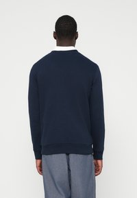 Bărbat în picioare, întors cu spatele, îmbrăcat cu un hanorac bleumarin, o cămașă albă dedesubt și pantaloni gri, pe un fundal simple.