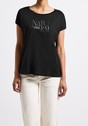 Person iført en sort kortærmet top med ordet "NATURAL" og sætningen "berøring giver liv til jorden," kombineret med hvide bukser.