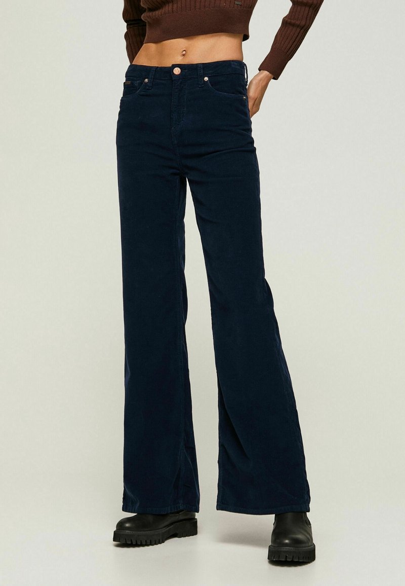 Femme portant un pantalon marine taille haute à jambes larges, des bottes noires épaisses et un pull court marron côtelé, debout avec une main dans la poche.