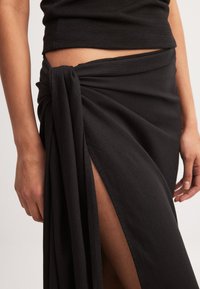 Juoda maxi sijonas, pagamintas iš minkštos medžiagos, su mazgo detalėmis juosmens srityje ir aukštu įpjovimu, suteikiančiu elegantišką išvaizdą. Lygi tekstūra ir lengvas dizainas.