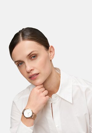 Goldfarbene Mesh-Armbanduhr mit einem runden weißen Ziffernblatt, das über silberne Stundenmarkierungen und eine Datumsanzeige verfügt, auf einer weißen Bluse.