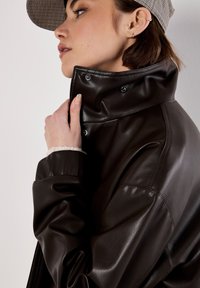 Braune Lederjacke mit hohem Kragen, Druckknöpfen und einem weichen, strukturierten Innenfutter, kombiniert mit einer Hahnentrittkappe.