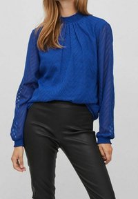 Blue long-sleeve blouse with a textured chevron pattern, gathered neckline, and elastic cuffs, paired with black fitted pants.