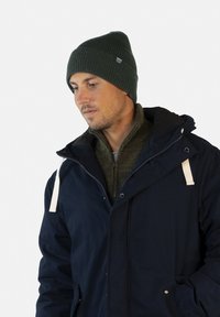 Parka bleu marine avec des accents de cordon blanc, portée par-dessus un pull vert ; arborant un bonnet côtelé vert foncé avec une étiquette de marque.