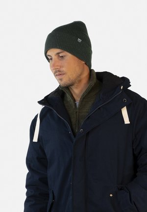 Parka bleu marine avec des accents de cordon blanc, portée par-dessus un pull vert ; arborant un bonnet côtelé vert foncé avec une étiquette de marque.