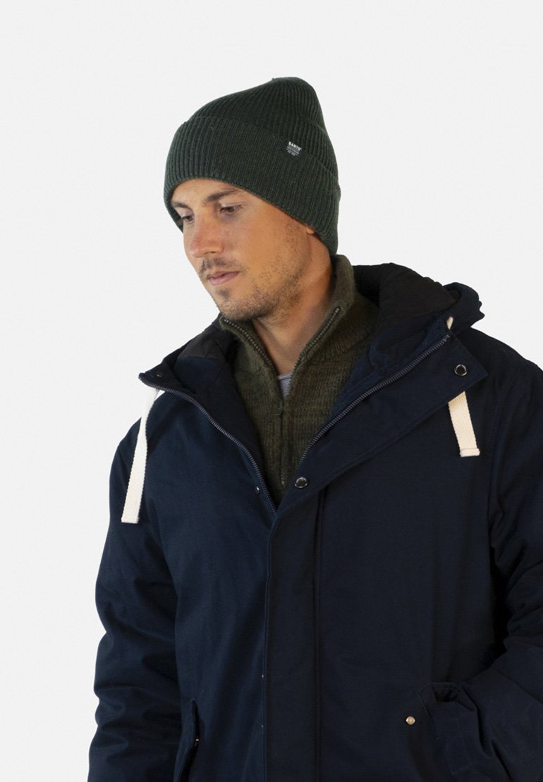 Parka bleu marine avec des accents de cordon blanc, portée par-dessus un pull vert ; arborant un bonnet côtelé vert foncé avec une étiquette de marque.