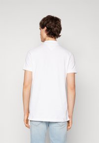 Polo blanco de textura suave, con mangas cortas, un cuello y un detalle en la parte posterior en forma de triángulo. Combina con unos pantalones vaqueros de color azul claro.