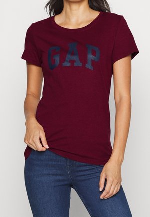 Kobieta w bordowej koszulce z krótkim rękawem z logo "GAP" i niebieskich dżinsach, trzymająca brzeg koszulki jedną ręką.