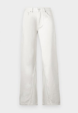 Personne en veste crème avec col noir et pantalon large crème tenant un sac à main noir, portant des chaussures noires, debout devant un arrière-plan uni.