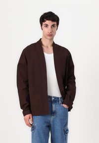 Brun, lett jakke med avslappet passform, åpen front og store lommer. Båret over en hvit t-skjorte og lyseblå jeans.