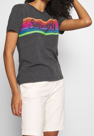 Personne porte un t-shirt gris foncé avec des rayures arc-en-ciel colorées et un texte en écriture cursive, assorti à un short blanc jusqu'aux genoux, sur fond blanc.