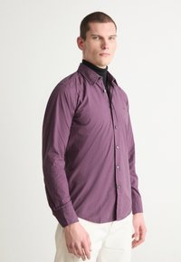 Langsleeve shirt in diep paarse stof met een button-down kraag, met een slim fit en contrasterende knopen. Gedragen over een zwarte coltrui.