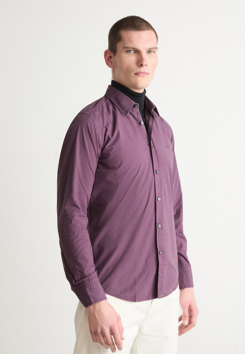Langsleeve shirt in diep paarse stof met een button-down kraag, met een slim fit en contrasterende knopen. Gedragen over een zwarte coltrui.