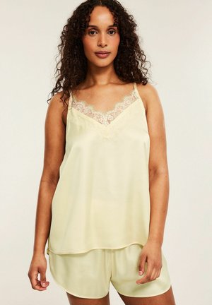 Frau trägt blassgelbes Satincamisole mit Spitzenbesatz und passenden Shorts, steht vor einfachem hellem Hintergrund.