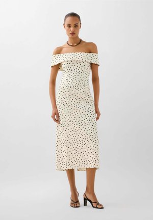 Mujer con un vestido midi blanco con hombros descubiertos y lunares negros, y sandalias negras de tacón, de pie frente a un fondo claro y liso.