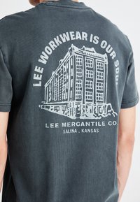 Ogle melnas t-kreklu ar vintage ēkās ilustrāciju un tekstu "LEE WORKWEAR IS OUR SOUL" zem kura ir "LEE MERCANTILE CO.".