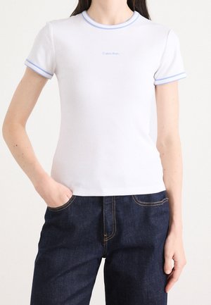 Kobieta ubrana w biały prążkowany top z krótkim rękawem i jasnoniebieskim obszyciem oraz małym logo Calvin Klein, w połączeniu z ciemnoniebieskimi dżinsami.