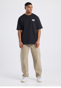 Black cotton t-shirt with a small white graphic on the left chest, paired with beige straight-leg pants and white sneakers.