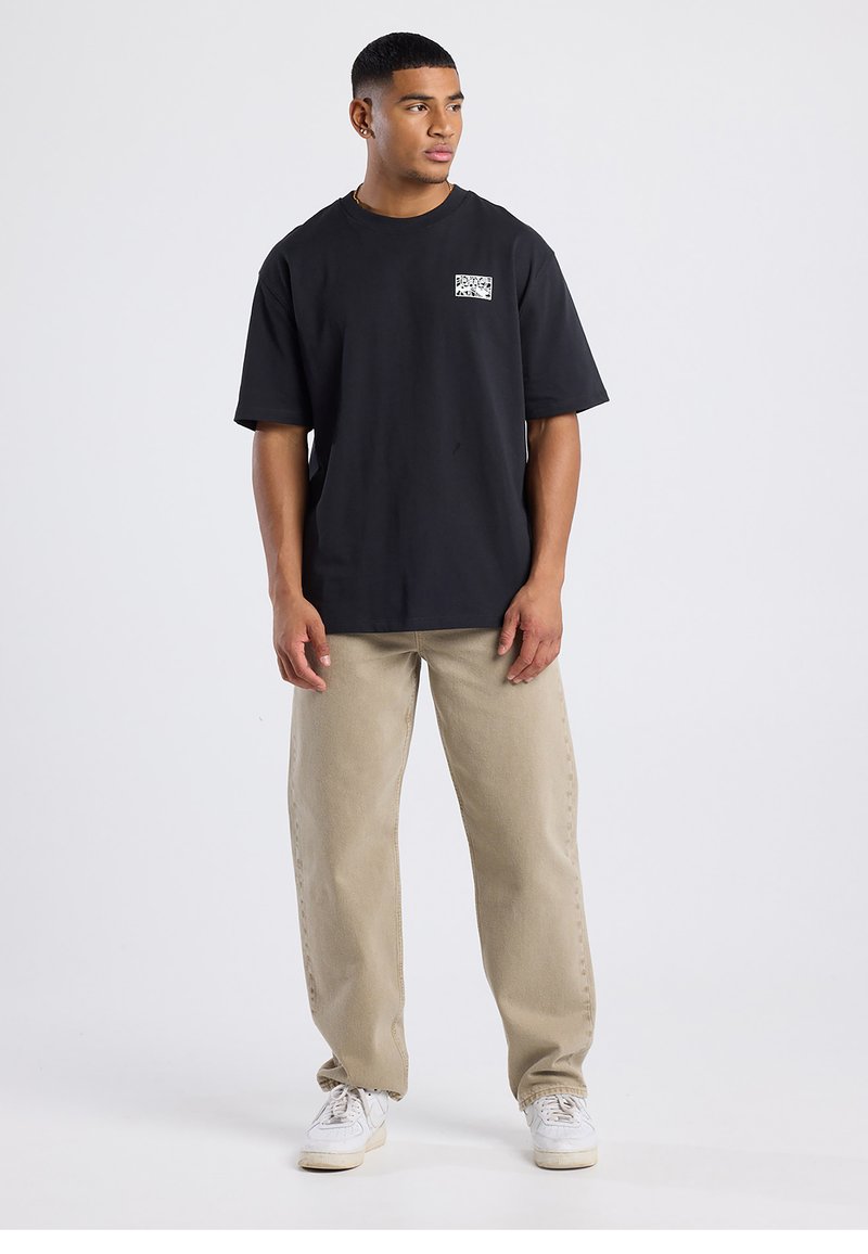 Black cotton t-shirt with a small white graphic on the left chest, paired with beige straight-leg pants and white sneakers.