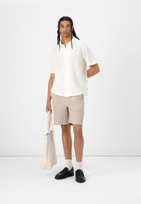 Vit kortärmad skjorta med ett strukturerat mönster, tillsammans med ljusbruna randiga shorts. Accessoarer inkluderar en beige tote-väska och svarta loafers.