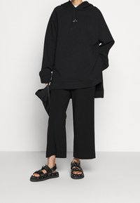 Felpa nera oversize con cappuccio e cordino, abbinata a pantaloni larghi neri e sandali neri con dettagli in fibbia e borchie.
