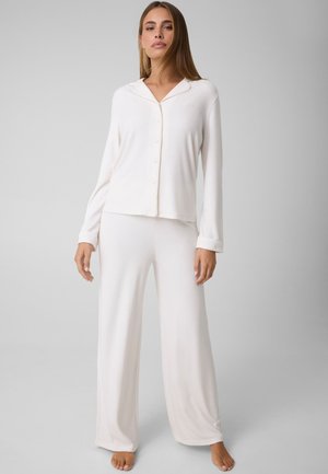 Femme debout pieds nus, portant un haut blanc à manches longues boutonné et un pantalon ample, sur un fond gris uni.