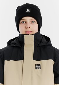 Schwarze und beige Skijacke mit hohem Kragen, Reißverschlüssen und auffälligem Logo auf der Brust. Begleitet von einer schwarzen Strickmütze.