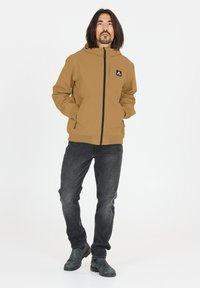 Brun huva jacka med dragkedja fram, elastiska ärmslut och sidofickor. Kombineras med mörka jeans och svarta skor. Enkel bakgrund.