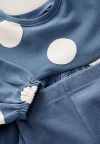 Pantalones de tela azul acanalada combinados con una camisa azul de manga larga con grandes lunares blancos y puños elásticos.