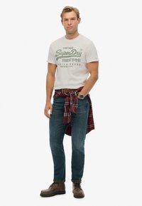 Conjunto masculino que incluye una camiseta de algodón blanca con estampado verde, jeans de mezclilla azul y botas de cuero marrón, completado con una camisa a cuadros roja atada a la cintura.