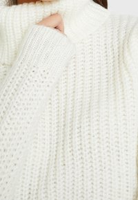 Chunky white knit sweater with a high, folded collar and ribbed texture. Features long sleeves and a relaxed fit. Soft, cozy material.