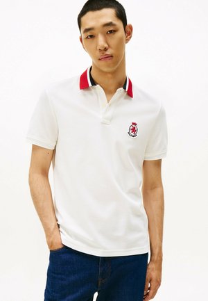 Hvid poloshirt med rød og marineblå krave, prydet med et broderet emblem på brystet. Stoffet har en struktureret overflade og korte ærmer.