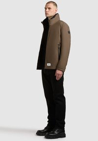 Brown insulated jacket with high collar, zip pockets, and black lining, worn over a black shirt and black pants, paired with chunky black shoes.