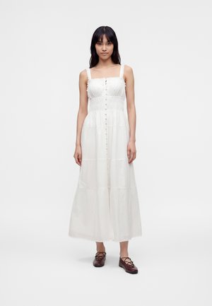 Femme aux longs cheveux foncés portant une robe midi blanche sans manches boutonnée et des chaussures plates marron foncé à lacets, debout sur un fond blanc.