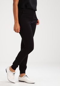 Femme portant un pantalon noir ajusté et des chaussures blanches à lacets, se tenant devant un fond blanc uni.