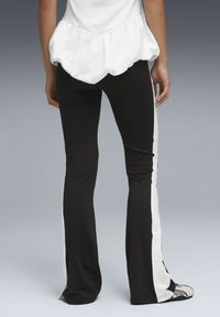 Άτομο που φοράει μαύρο παντελόνι με λευκές πλαϊνές ρίγες και κουμπιά, σε συνδυασμό με λευκό τοπ, στέκεται μπροστά σε γκρι φόντο.