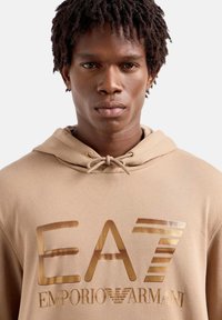 Felpa con cappuccio beige caratterizzata da un grande logo metallico oro "EA7" e il testo "Emporio Armani". Tessuto liscio con una vestibilità comoda.